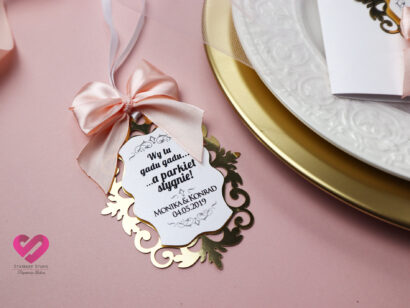 Pastelowe, romantyczne zawieszki ślubne na alkohol w stylu glamour z delikatną różową wstążką i złotym ażurem pasujące do zaproszeń