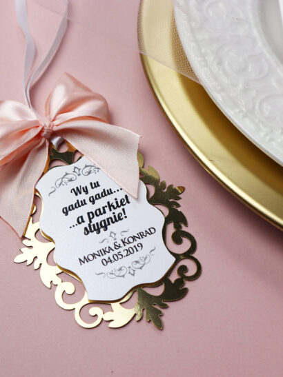 Pastelowe, romantyczne zawieszki ślubne na alkohol w stylu glamour z delikatną różową wstążką i złotym ażurem pasujące do zaproszeń